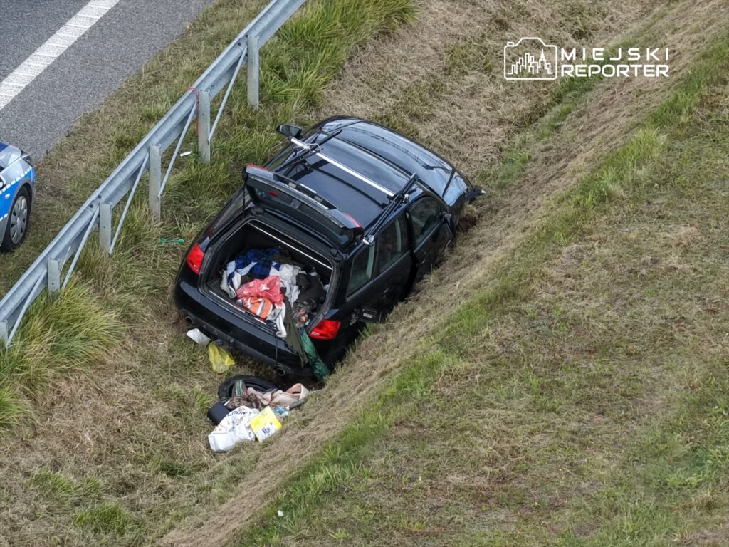 Czarny samochód osobowy w rowie przy drodze, otwarty bagażnik z widocznymi ubraniami i odpadkami. Obok policyjny radiowóz.