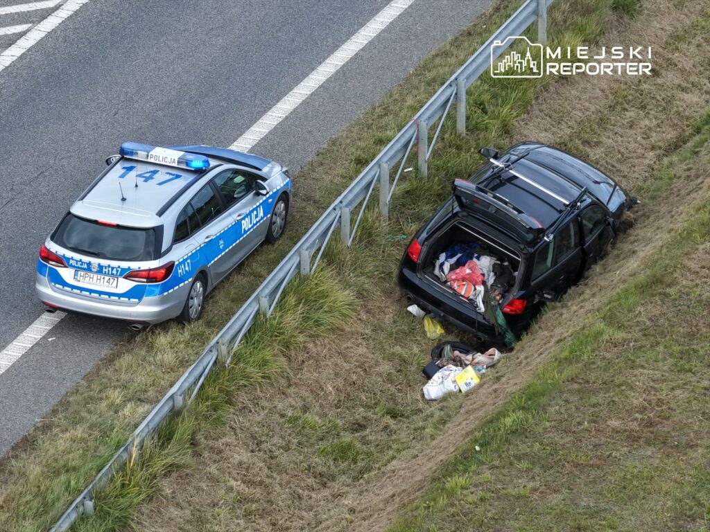Policyjny radiowóz zatrzymany przy poboczu drogi obok czarnego samochodu z otwartą klapą bagażnika, wypełnionym odpadkami.
