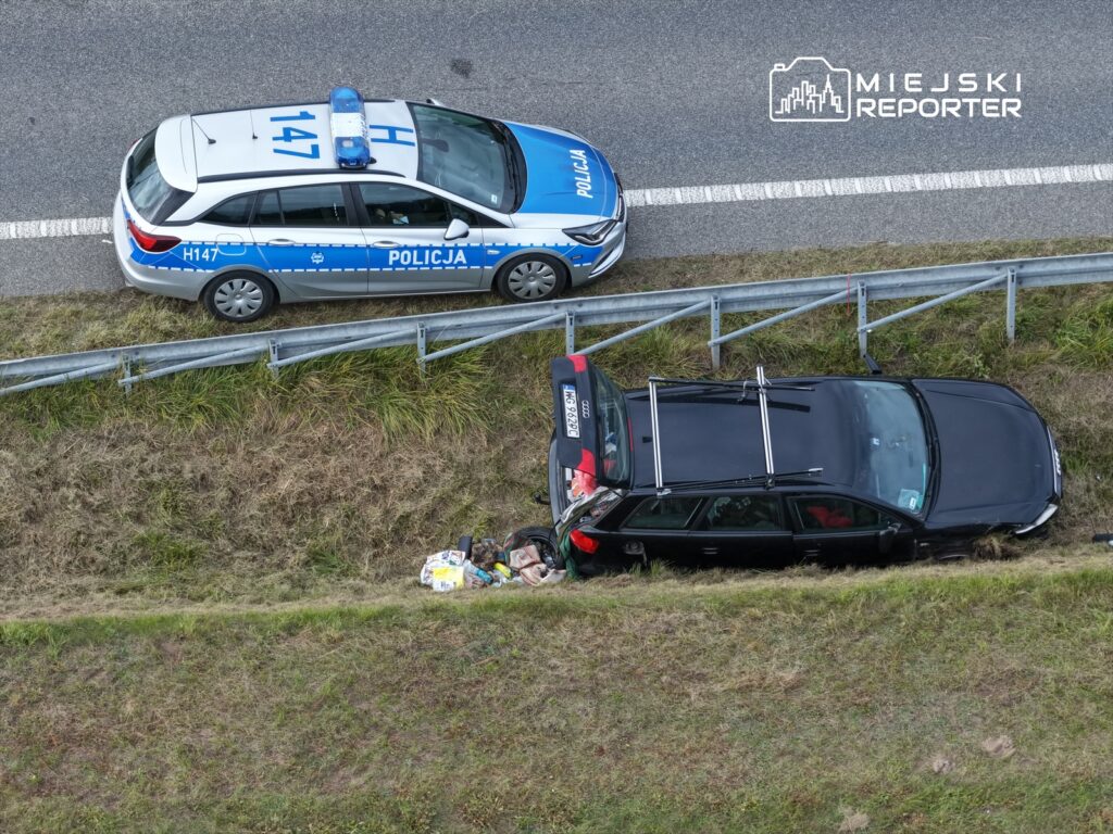 Radiowóz policji zatrzymany przy poboczu drogi obok uszkodzonego czarnego samochodu osobowego w rowie.