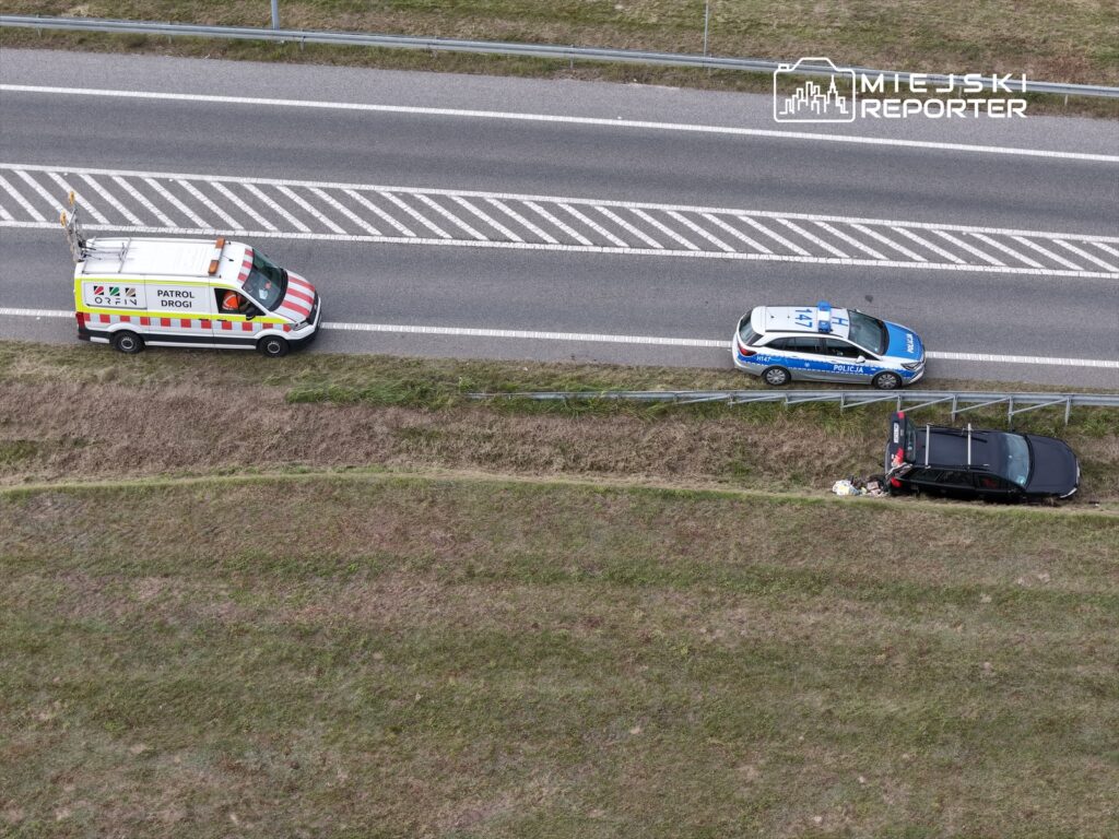 Na drodze zatrzymane są pojazdy: samochód policyjny oraz furgonetka patrolu drogowego obok uszkodzonego auta.