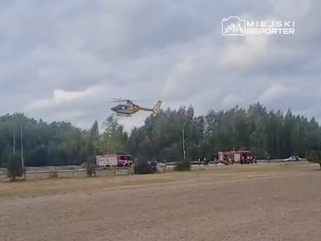Helikopter ratunkowy przelatuje nad terenem z pojazdami straży pożarnej w tle, otoczony drzewami.