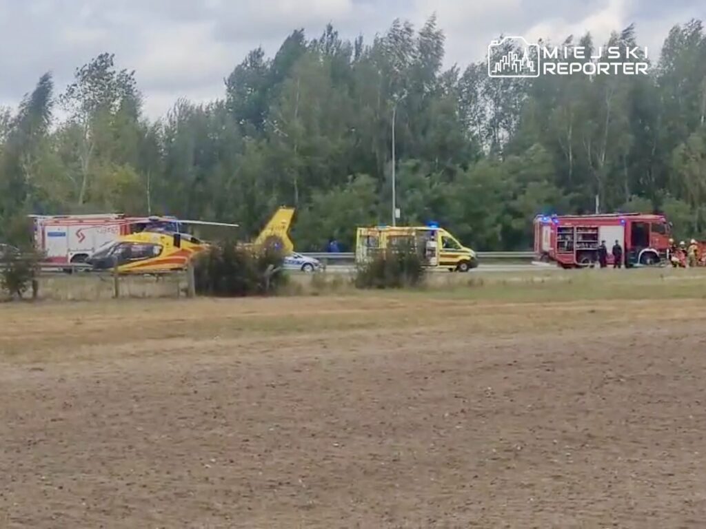 Na drodze stoją karetki, wóz strażacki oraz śmigłowiec ratunkowy, a w tle widać zalesiony teren.