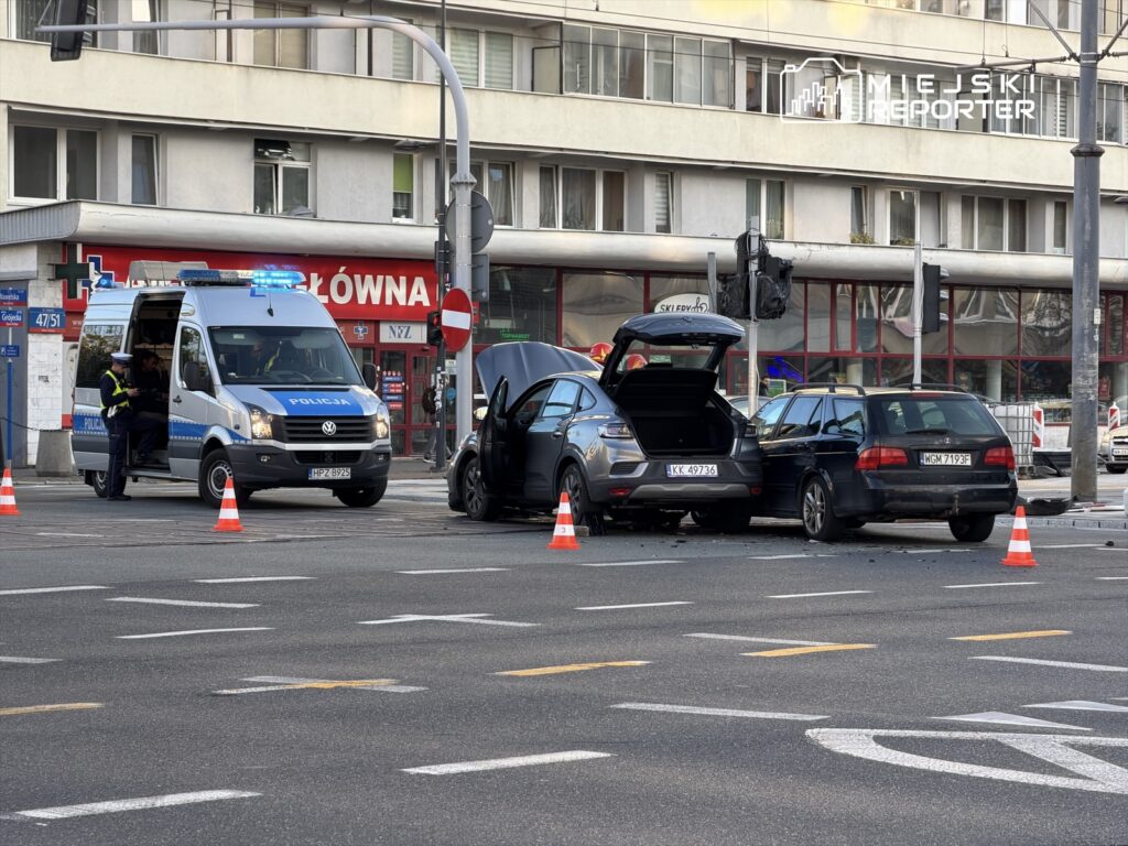 Na skrzyżowaniu stoją dwa uszkodzone samochody, wokół których ustawione są pachołki. Policjant rozmawia z osobami w policyjnym radiowozie.