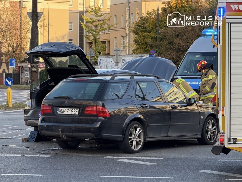 Czarny samochód osobowy z otwartą klapą bagażnika stoi na skrzyżowaniu, obok niego pracują strażacy.
