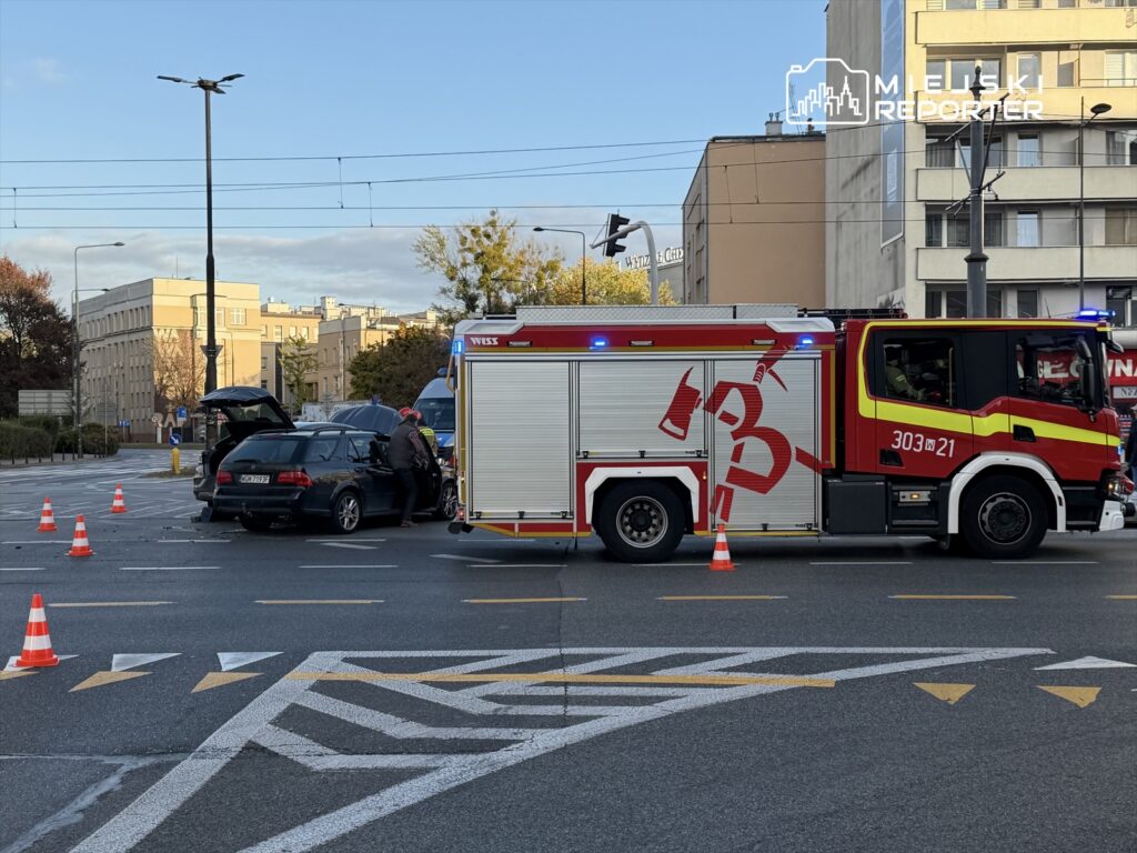 Strażacy pracują nad usunięciem skutków wypadku drogowego na skrzyżowaniu, obok zaparkowanego samochodu.