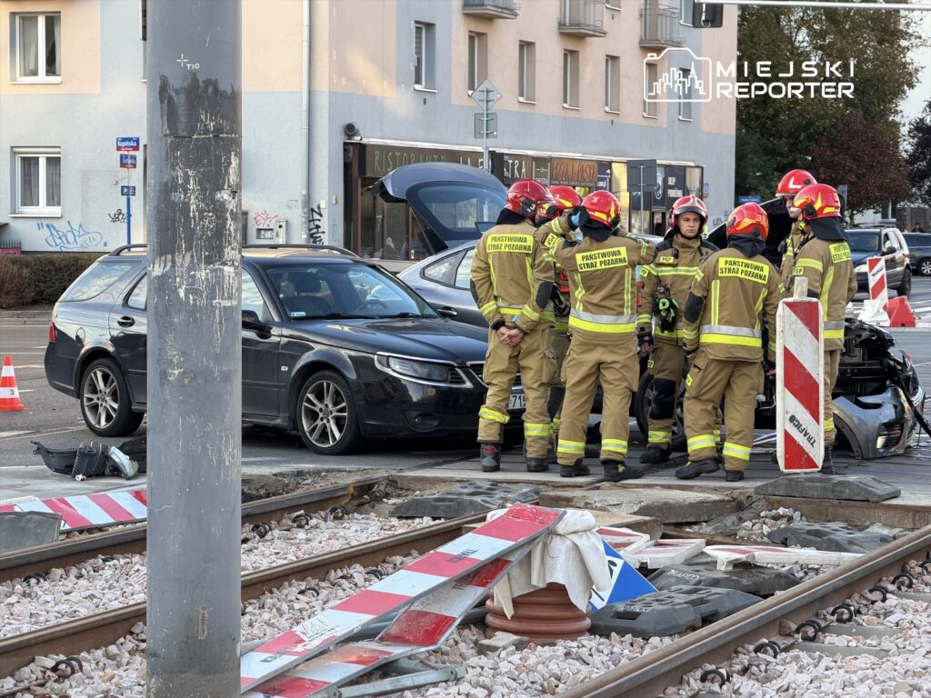 Grupa strażaków w mundurach rozmawia obok uszkodzonego samochodu przy torach kolejowych.
