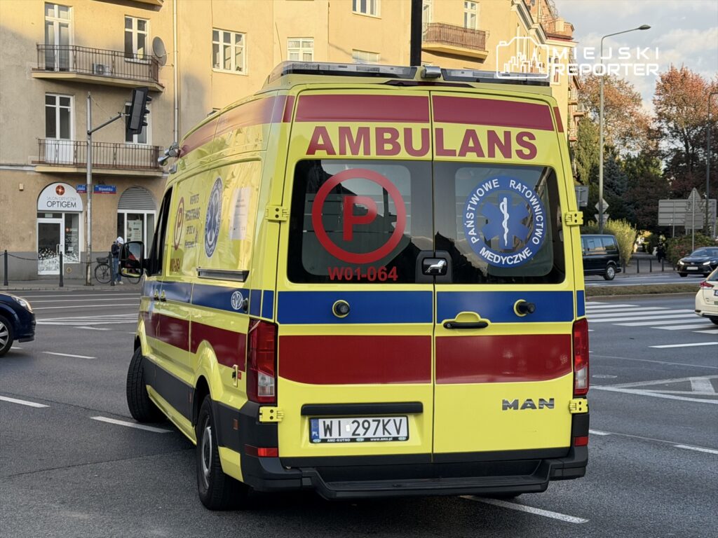 Ambulans z widocznym napisem "AMBULANS" zatrzymany na skrzyżowaniu, w tle budynki mieszkalne i sklepy.