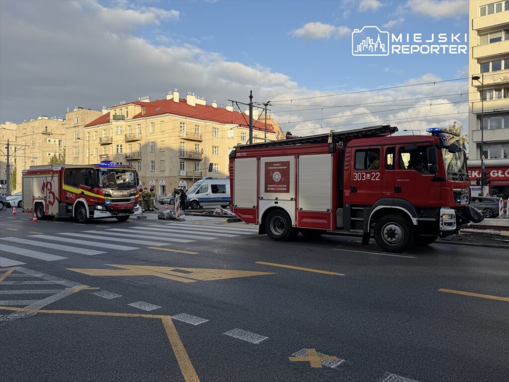 Dwa wozy strażackie na ulicy, jeden z włączonymi sygnałami, obok ekipa ratunkowa pracująca przy drodze.