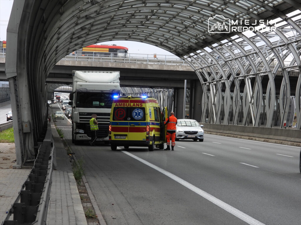 Ambulans z włączonymi sygnałami zatrzymany obok ciężarówki na drodze, obok pracownik w kamizelce odblaskowej.
