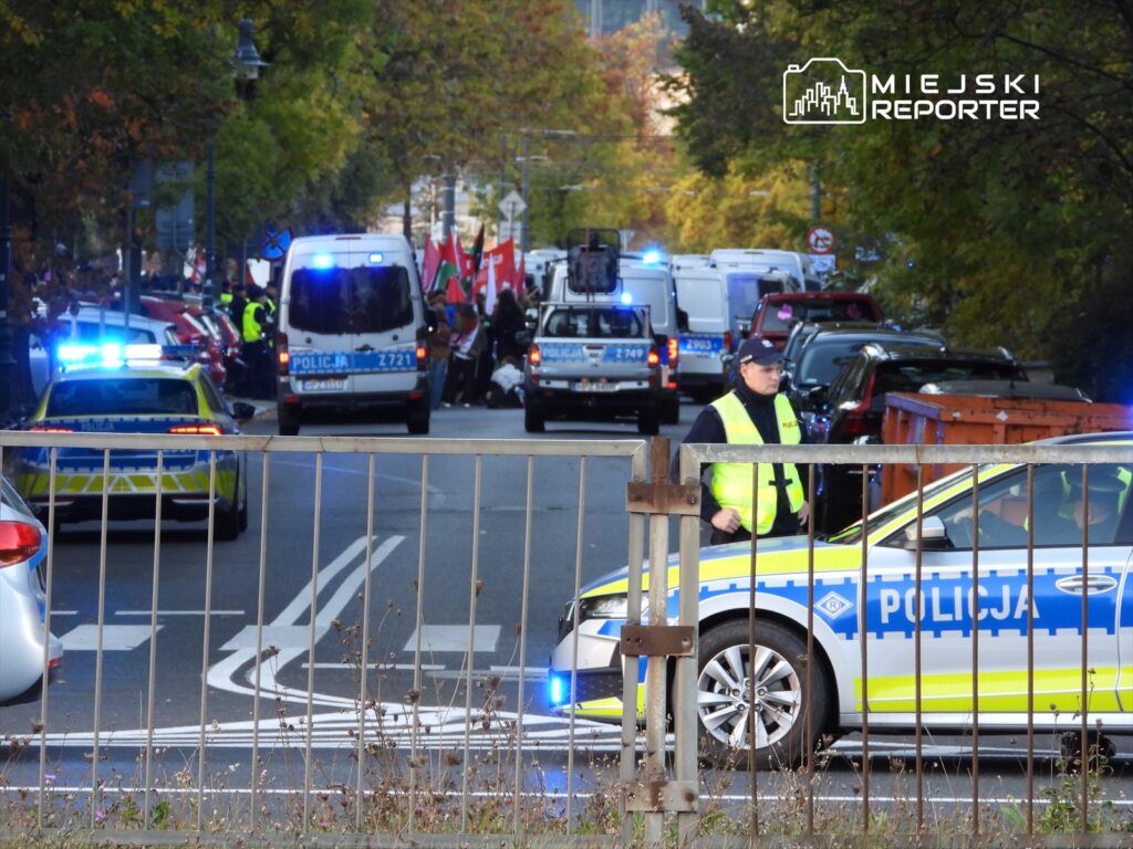 Policyjny radiowóz z włączonymi sygnałami świetlnymi stoi na ulicy, a funkcjonariusz w kamizelce odblaskowej rozmawia z innymi policjantami. W tle widać tłum ludzi z flagami.