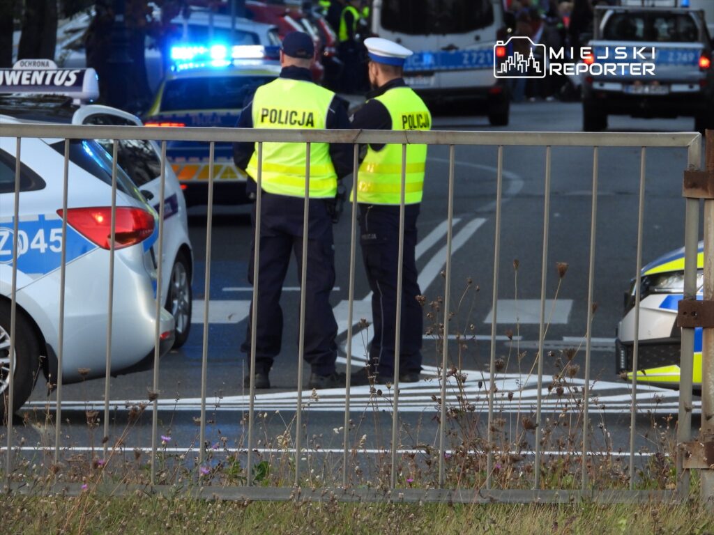 Dwaj policjanci w kamizelkach odblaskowych rozmawiają przy zablokowanej ulicy, obok radiowozów z włączonymi sygnałami.