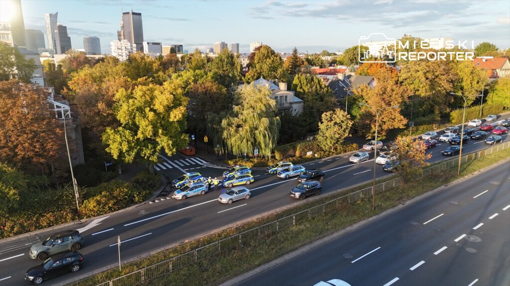 Grupa radiowozów policyjnych zatrzymuje ruch na ulicy, otoczonej drzewami w jesiennych barwach.