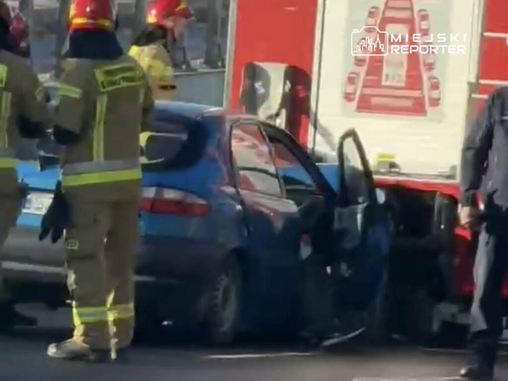 Grupa strażaków w mundurach ocenia uszkodzone niebieskie auto po wypadku na ruchliwej drodze.