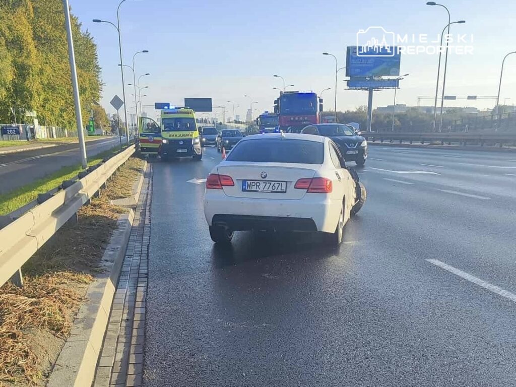 Biały samochód osobowy zatrzymany na pasie ruchu, obok stoją pojazdy służb ratunkowych z włączonymi sygnałami świetlnymi.