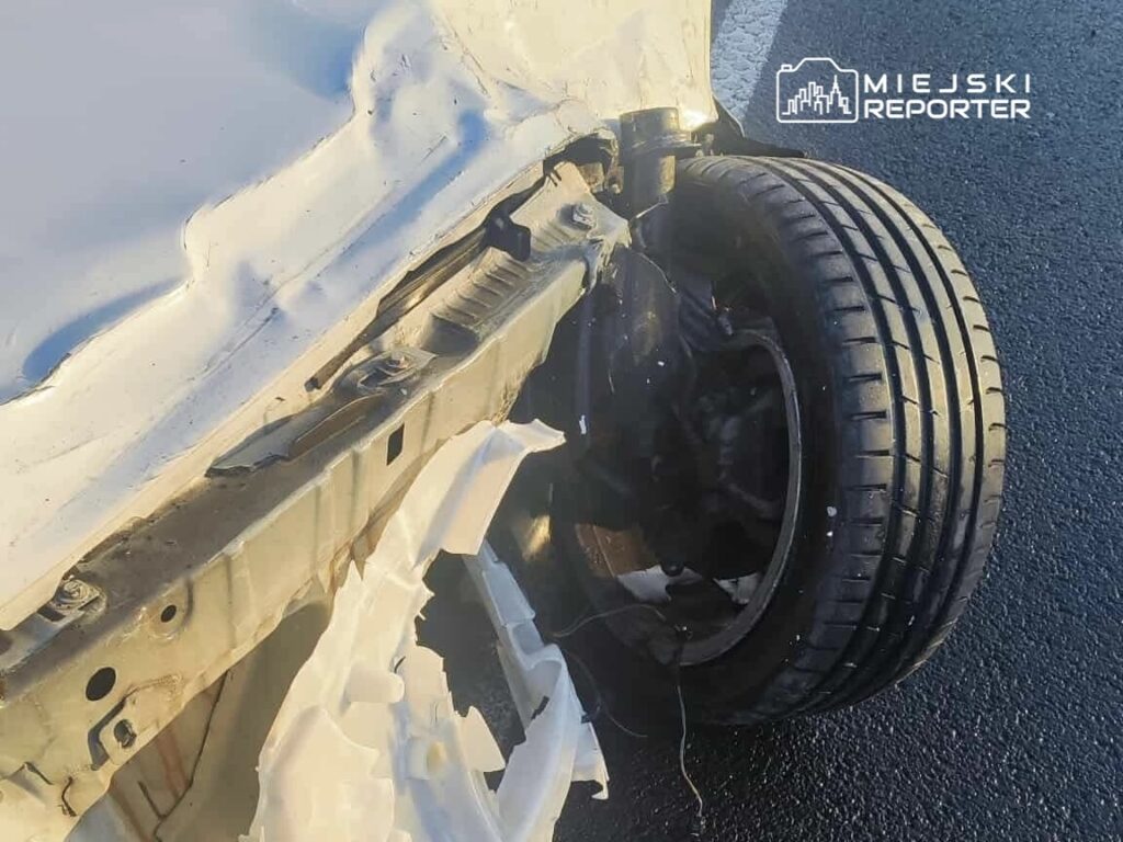 Uszkodzony pojazd z odgiętym nadwoziem i widoczną oponą leżący na drodze.