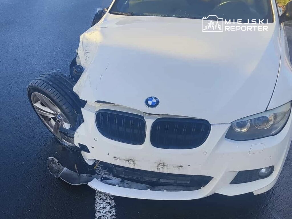 Uszkodzony biały samochód marki BMW z zniszczonym przodem i oderwaną oponą na asfaltowej drodze.