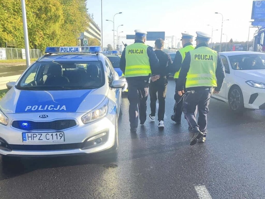 Czterech policjantów w kamizelkach odblaskowych prowadzi zatrzymanego mężczyznę na ruchliwej ulicy.