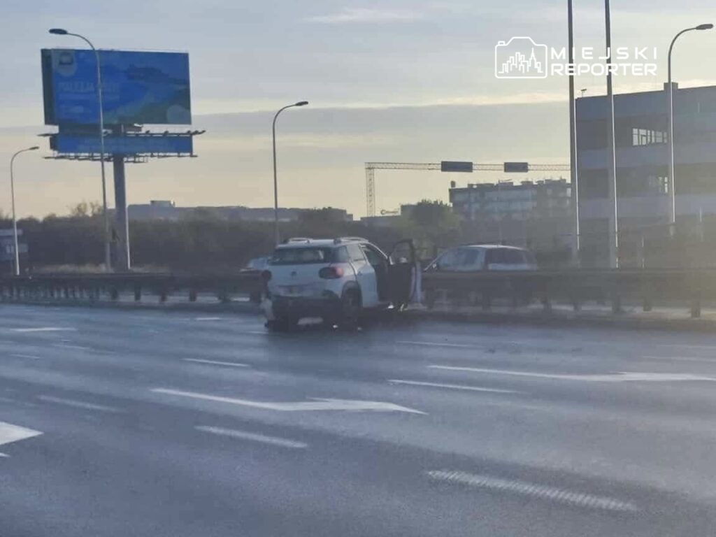 Samochód osobowy zatrzymany na pasie drogi, z otwartymi drzwiami, obok inny pojazd, w tle billboard reklamowy.