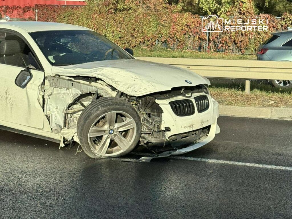 Zniszczony samochód osobowy marki BMW z uszkodzoną przednią częścią stoi na drodze obok ogrodzenia.