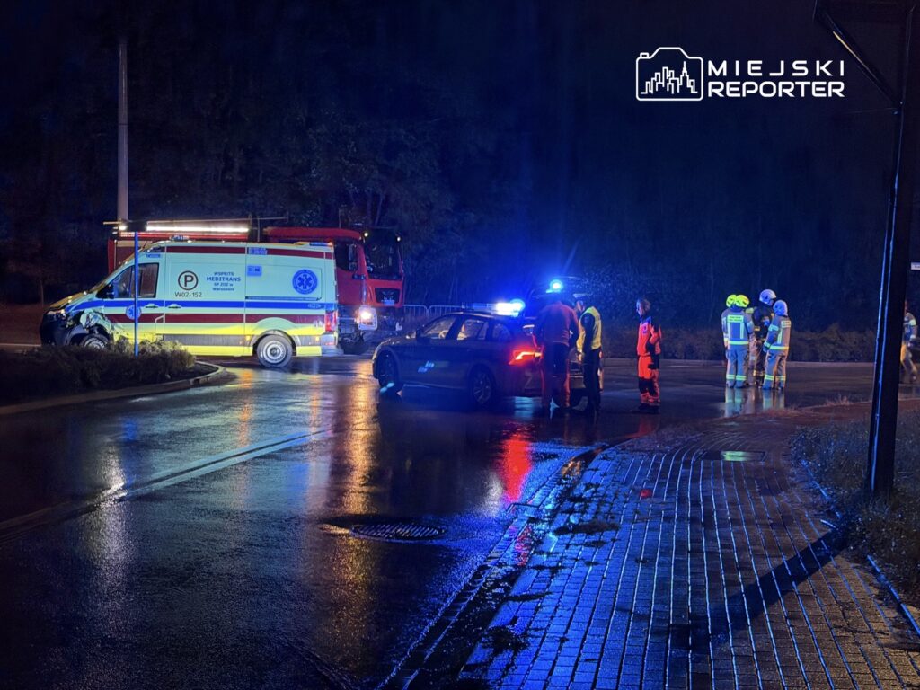 Zespół ratunkowy w odblaskowych kamizelkach rozmawia obok wozu strażackiego na mokrej drodze nocą.