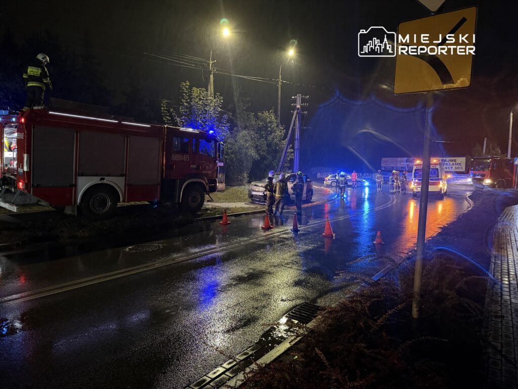 Strażacy pracują przy wypadku drogowym w deszczową noc, otoczeni przez pojazdy i sygnalizację świetlną.