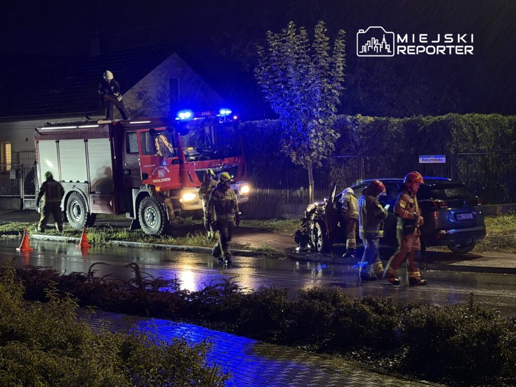 Strażacy w mundurach badają uszkodzony samochód na oświetlonej ulicy w nocy, obok stoi wóz strażacki.