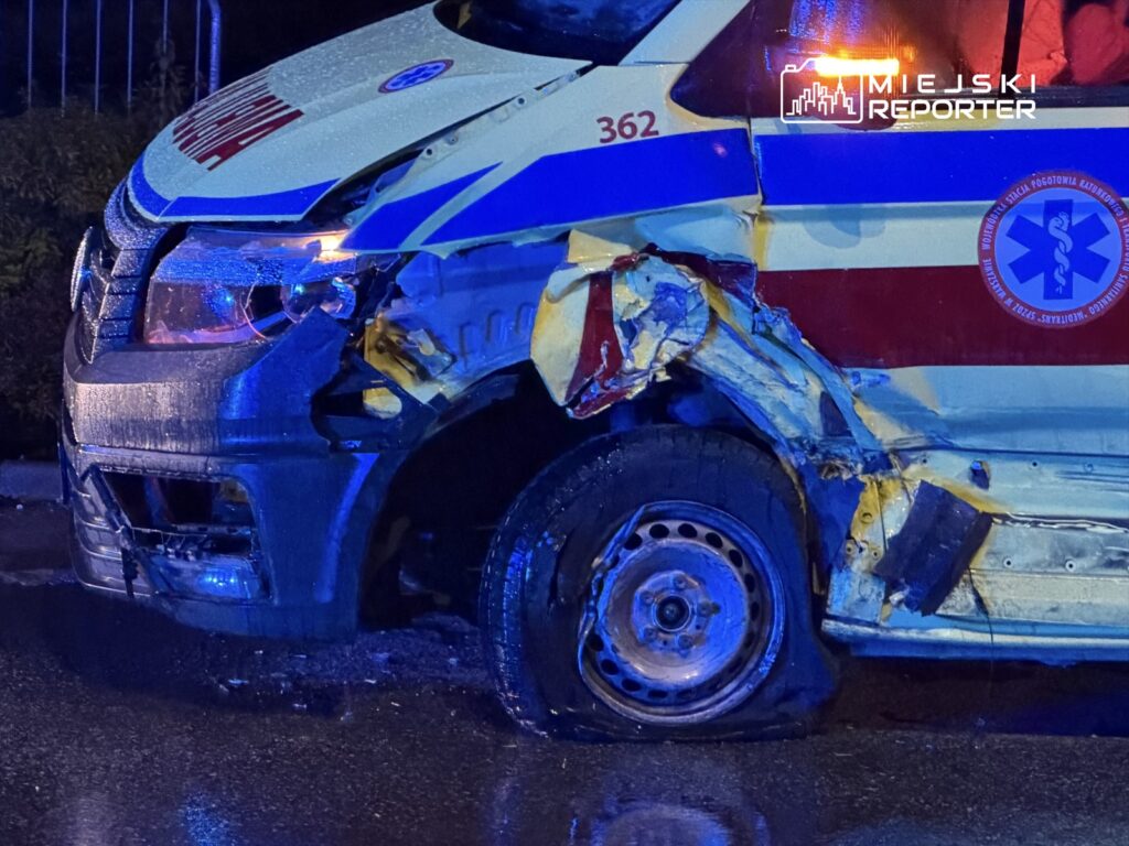 Uszkodzony ambulans z zniszczoną przednią częścią, stojący na mokrej nawierzchni w nocy, oświetlony niebieskimi światłami.