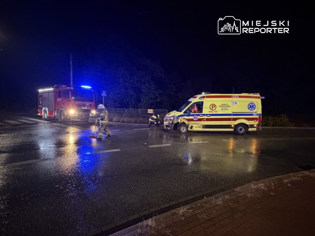 Strażacy usuwają wodę z jezdni po wypadku drogowym, obok stoi woz strażacki oraz ambulans w nocnym otoczeniu.