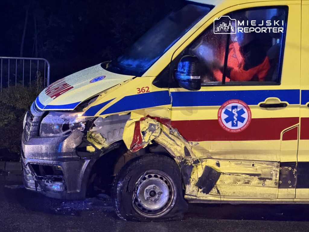 Uszkodzony ambulans z wgniecioną boczną częścią i zerwaną oponą, zaparkowany w ciemnej okolicy po deszczu.