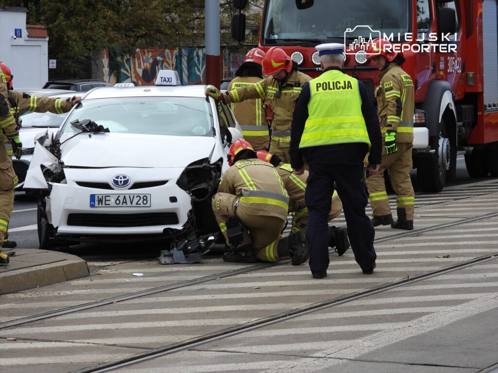 Grupa strażaków oraz policjant w kamizelce odblaskowej badają uszkodzony samochód osobowy na skrzyżowaniu.