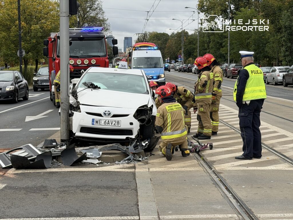 Biały samochód osobowy uderzył w słup, a strażacy oraz policja udzielają pomocy na miejscu zdarzenia.