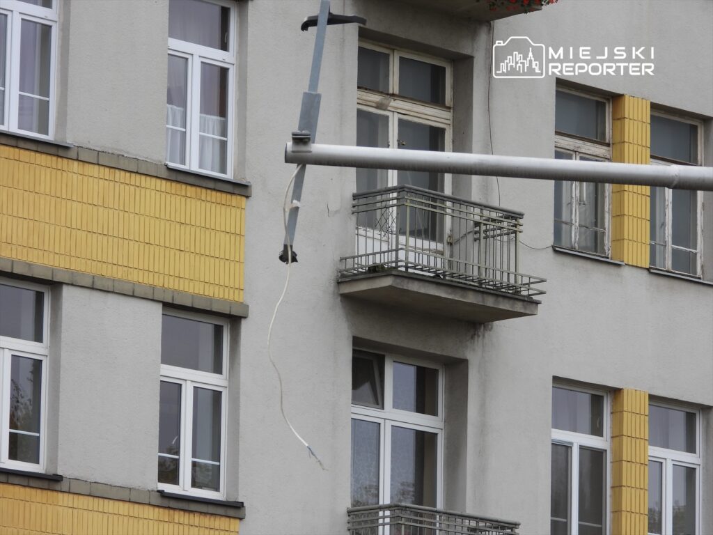 Zawieszony kabel elektryczny przy budynku mieszkalnym z żółtymi elementami, obok balkon z metalową balustradą.