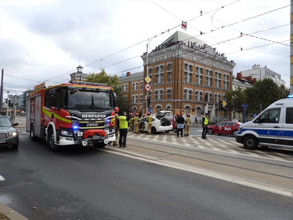 Straż pożarna i policja obsługują wypadek drogowy na skrzyżowaniu, w tle zabytkowy budynek z czerwonymi detalami.