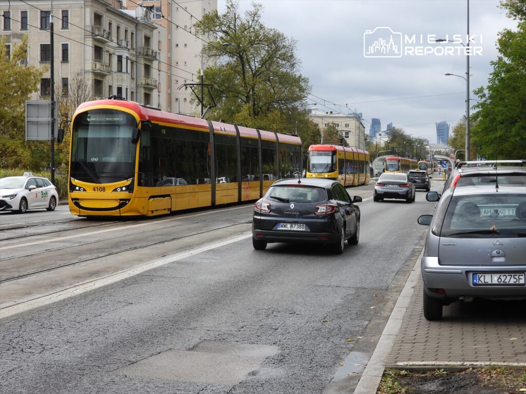 Tramwaj w żółto-czerwonym malowaniu przejeżdża obok zaparkowanych samochodów na miejskiej ulicy.