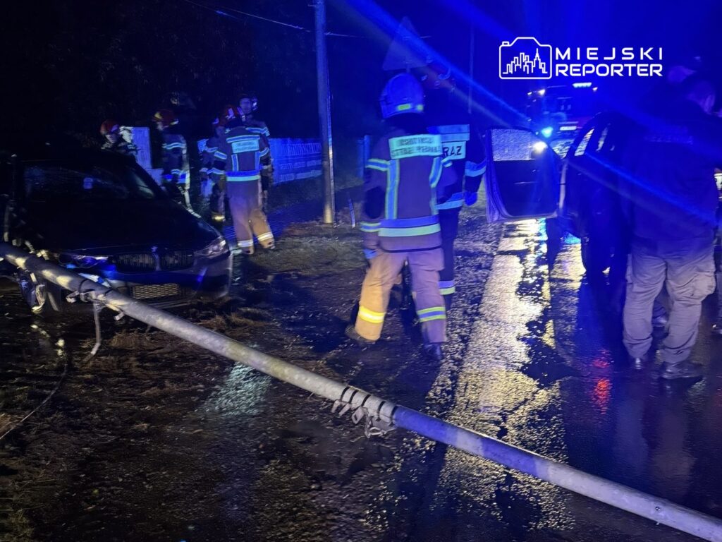 Grupa strażaków w mundurach z odblaskowymi paskami pracuje przy uszkodzonym samochodzie obok przewróconego słupa energetycznego.