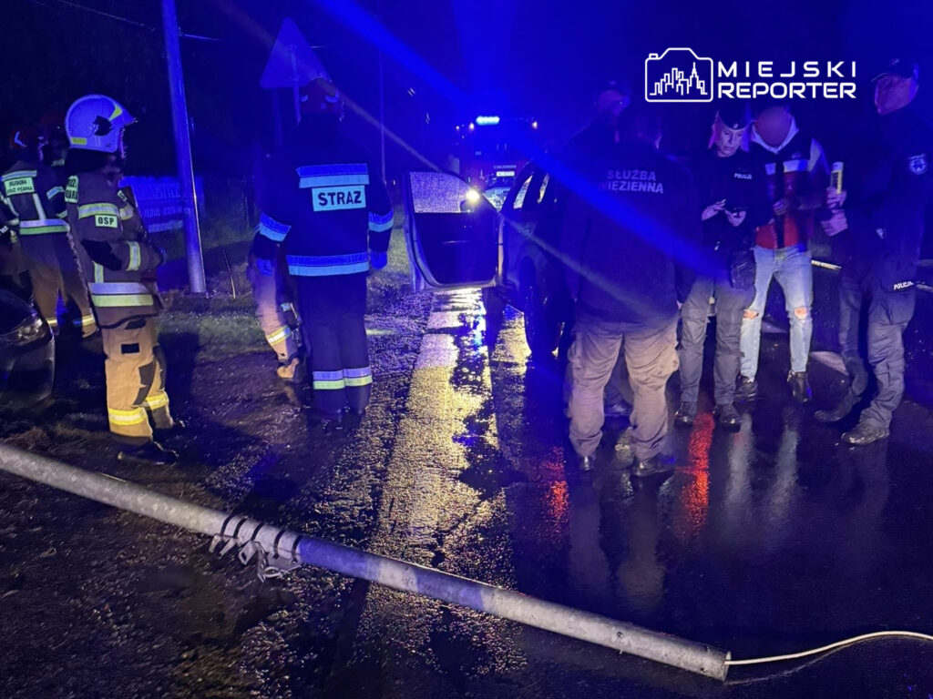 Grupa strażaków i policjantów zbiera się wokół uszkodzonego pojazdu na mokrej drodze, obok leżącego słupa.