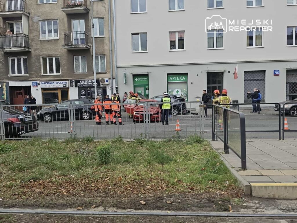 Grupa ratowników i policjantów pracuje na miejscu wypadku drogowego przy ulicy, obok zaparkowanych samochodów.