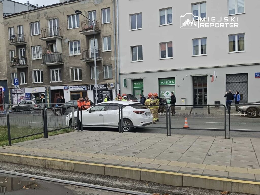 Grupa ratowników w pomarańczowych kamizelkach pracuje przy białym samochodzie na ulicy obok apteki.
