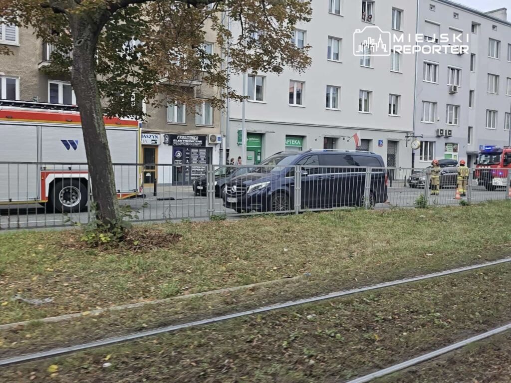 Strażacy z wozem gaśniczym pracują przy ul. z widocznymi pojazdami i budynkami w tle.