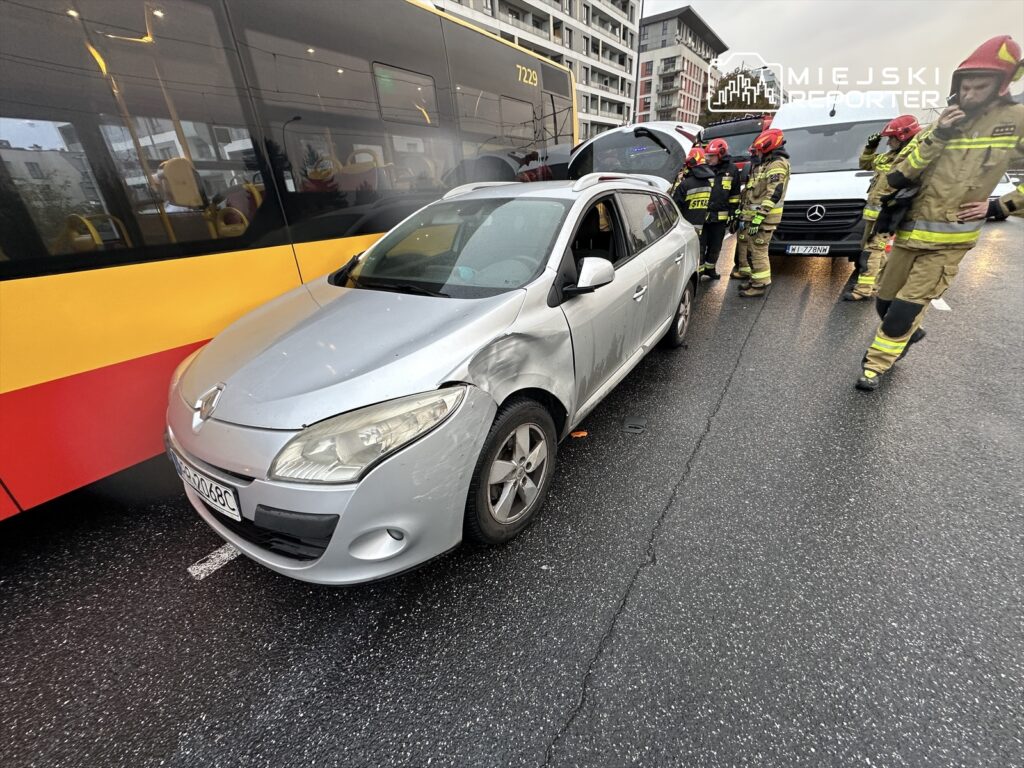 Srebrny samochód osobowy z uszkodzoną przednią częścią stoi obok autobusu, a obok niego pracują strażacy w mundurach.