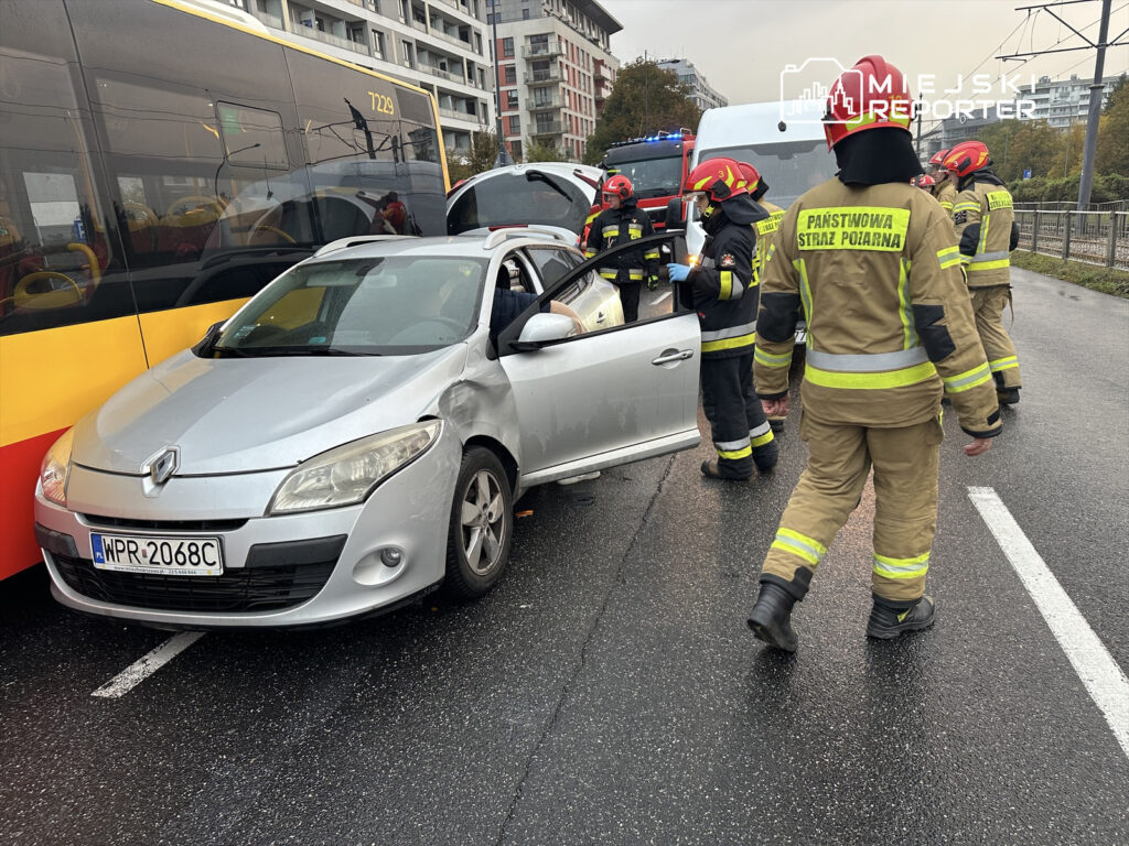 Srebrny samochód osobowy z uszkodzoną stroną stoi obok autobusu, a strażacy w mundurach przeprowadzają akcję ratunkową.