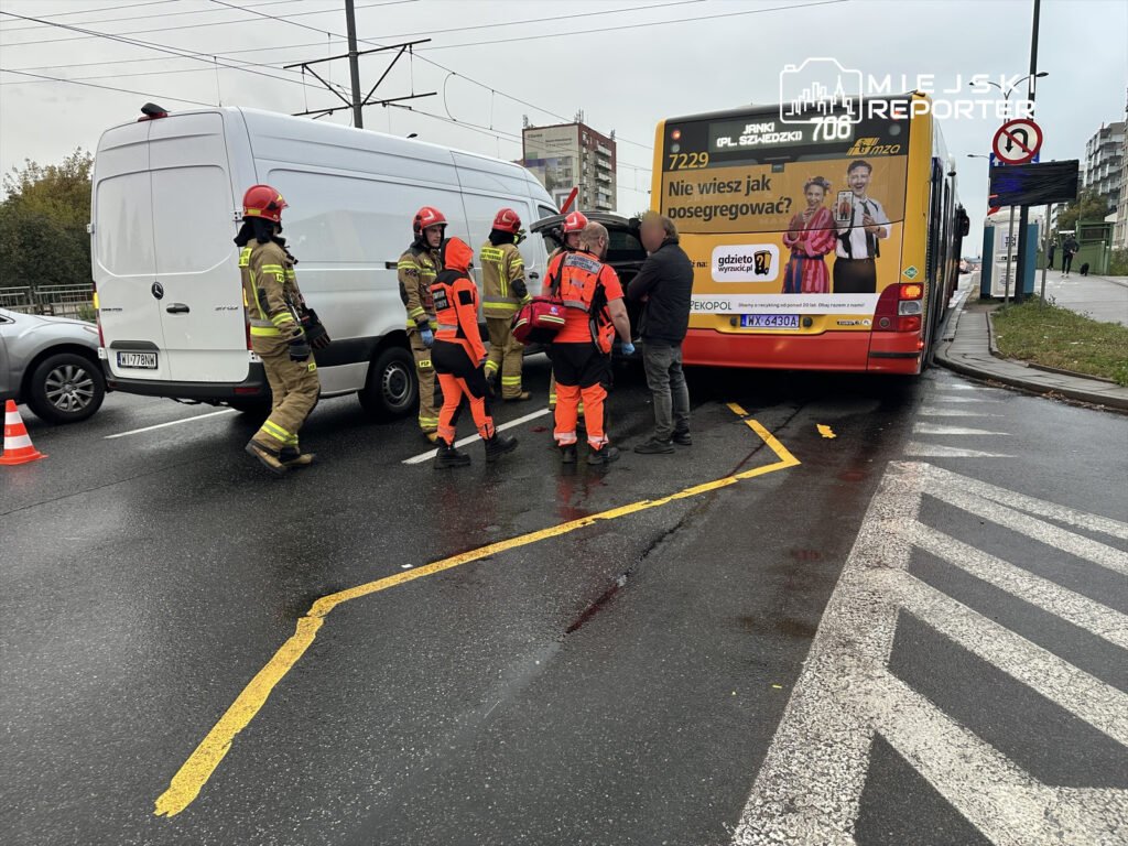 Grupa strażaków i ratowników w odzieży ochronnej pracuje przy zderzeniu autobusu z samochodem dostawczym na ruchliwej ulicy.