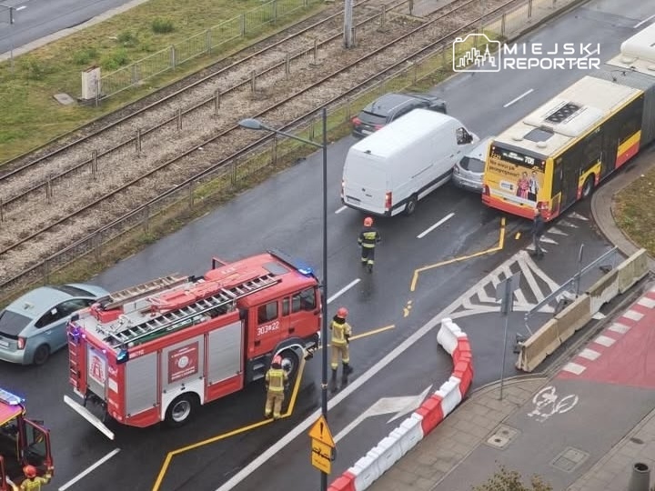 Na skrzyżowaniu ulicy z torami stoi wóz strażacki, a obok pracują strażacy przy zderzeniu trzech pojazdów.