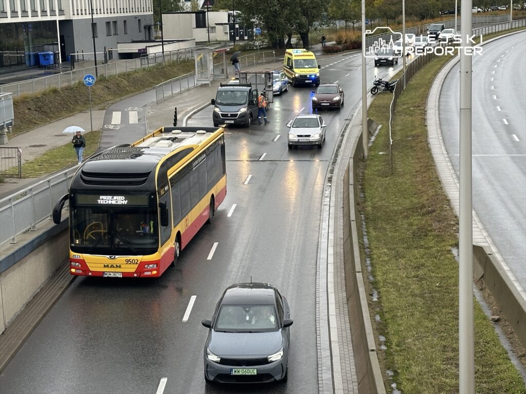 Żółto-czerwony autobus miejskie zatrzymany na przystanku, obok ruchliwa droga z samochodami i karetką.
