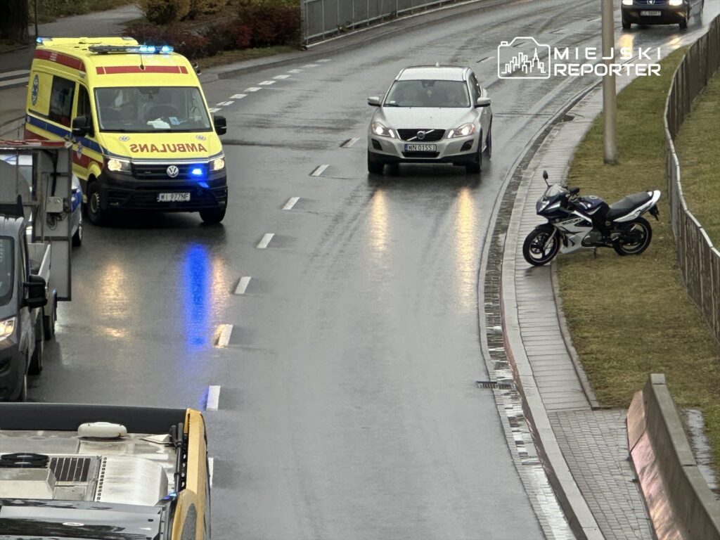 Ambulans z włączonymi sygnałami świetlnymi zatrzymany na mokrej drodze, obok zaparkowany motocykl i przejeżdżający samochód.