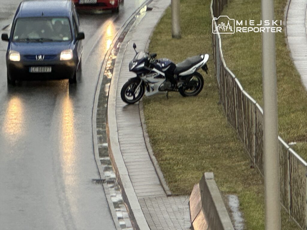 Motocykl zaparkowany na trawie obok jezdni, obok przejeżdżające samochody w deszczowej aurze.