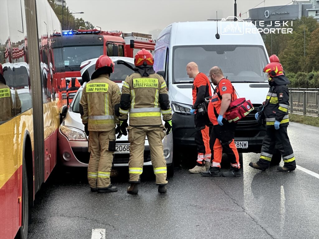 Grupa strażaków i ratowników medycznych udziela pomocy na miejscu wypadku, gdzie samochód osobowy jest zgnieciony między dwoma pojazdami.