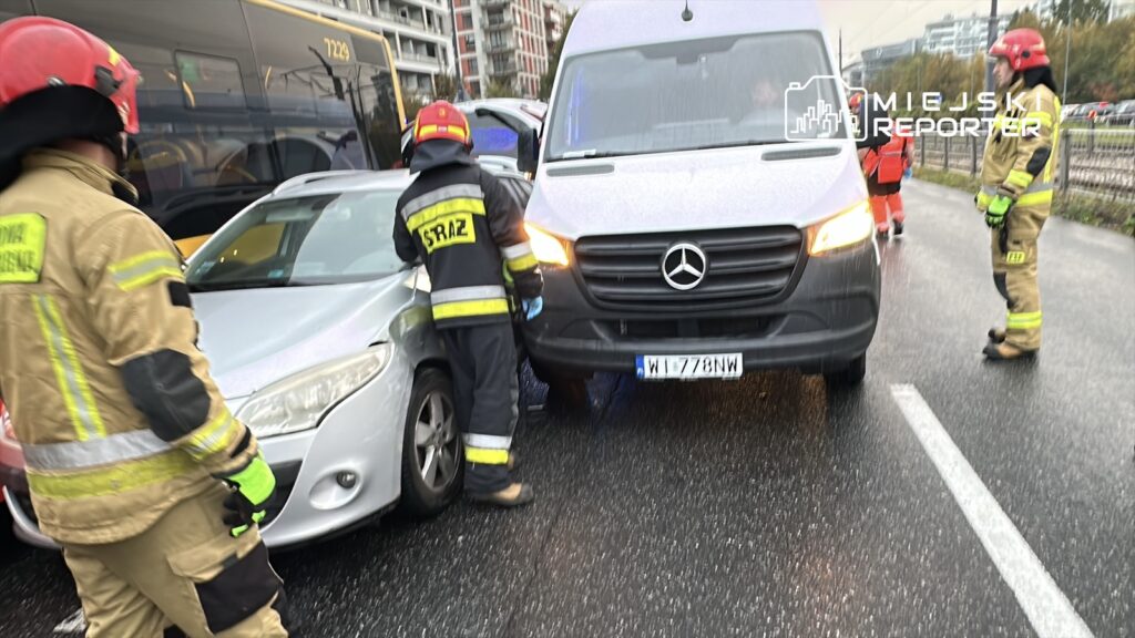 Strażacy w mundurach udzielają pomocy po kolizji dwóch pojazdów na deszczowej ulicy.
