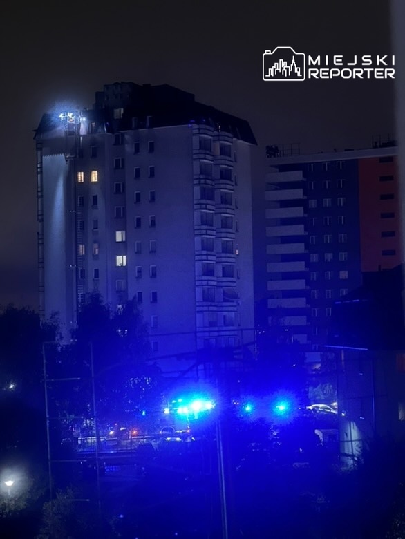 Nocny widok na budynek mieszkalny z niebieskimi światłami radiowozów w tle oraz reflektorem na dachu.