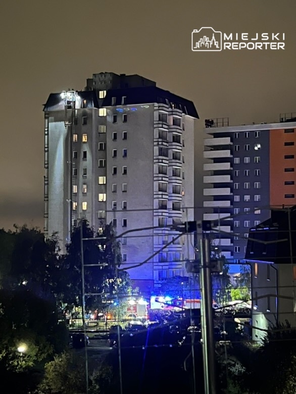 Straż pożarna z wozami ratunkowymi interweniuje przy wysokim budynku mieszkalnym w nocy, w tle widoczne światła.